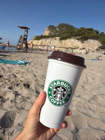 A hand holds a white Starbucks coffee cup with a brown lid in the foreground. The cup has the Starbucks logo printed in green and white. The background depicts a sandy beach under a clear blue sky. Several people are visible on the beach, some near the water and others spread out on the sand. A wooden lifeguard stand is situated further back on the beach, and a rocky, tree-covered hill rises behind it. The scene appears to be set in Urbanització Cales I Talaies, Spain, based on the visual cues of a Mediterranean beach environment.