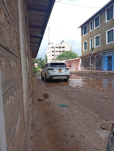 The image displays an unpaved, muddy street in Shella ward, Kenya, under an overcast sky. A white SUV, license plate KDC 111G, is positioned with its rear facing the viewer, partially submerged in a large water puddle centrally located in the roadway. To the left, a multi-story building constructed from rough stone blocks with a corrugated metal roof overhang extends along the frame. To the right, a three-story building with a light-colored, textured facade features multiple windows with dark frames and several identical wooden doors at ground level. One door is open, revealing a person seated inside wearing a light-colored garment. A sign above an adjacent closed door reads "OFFICE." Beyond this structure, a smaller, single-story pink building is visible. In the background, a tall, multi-story white building with two communication antennas on its roof rises above a large green tree. The ground surface is uneven, consisting of dirt and gravel, heavily saturated with water, forming numerous reflective puddles. Debris, including several rocks and a piece of tangled green material, is scattered on the ground. A portion of a person's leg and foot, wearing patterned clothing, is visible in the bottom right corner.
