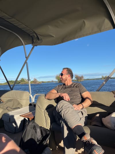 A man wearing sunglasses, a dark gray t-shirt, and light gray shorts is sitting on a boat. The boat appears to be moving on a wide river, possibly the Zambezi River, based on the location provided. The man is partially reclined on the boat's seating. A large, dark backpack is nearby, as well as a wooden tray or cutting board. The boat has a canopy overhead, shielding some of the view of the clear blue sky. The riverbank is visible in the background.