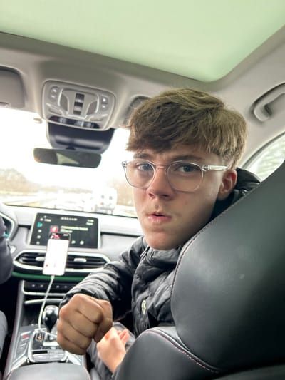 The scene depicts the interior of a modern vehicle traveling on a multi-lane road in Steinbourg, France, viewed from a rear passenger perspective. A young male individual, with light brown hair and clear-framed glasses, occupies the front passenger seat, looking directly towards the camera. He wears a black puffer jacket. His mouth is slightly agape, and his right hand is clenched into a fist, resting near the center console. A white smartphone is vertically mounted on the dashboard, connected by a white cable, displaying an illuminated interface. The vehicle's infotainment screen is active, showing various icons and what appears to be a navigation or media display. The overhead console with controls is visible, and the vehicle has a panoramic sunroof. Outside the vehicle, other vehicles, including a white recreational vehicle, are visible on the road under an overcast sky, with indistinct roadside foliage.