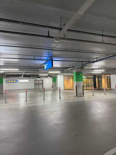This is a photograph of a parking garage located at the Lotte Outlets and IKEA in Goyang-si, South Korea. The scene is illuminated by overhead fluorescent lights, casting a uniform glow on the concrete floor and ceiling. Several metal pipes and electrical conduits are visible running along the ceiling.
In the background, signs indicate "IKEA" and "LOTTE OUTLETS," with an "Entrance" sign above double doors. Pillars, some painted bright green, support the structure. Bolted to the floor at regular intervals are silver bollards topped with red and white reflective bands. The floor is a smooth, gray concrete surface with white painted lines marking parking bays or lanes. The overall impression is one of a functional, modern indoor public space. There are no people visible in the frame.
