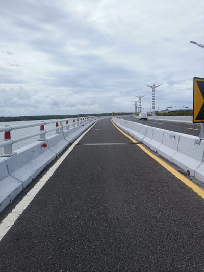 A long, empty asphalt road curves to the right, bordered by a white concrete barrier topped with a white railing and red and white posts. The railing is parallel to a body of water on the left, with a treeline visible in the distance. On the right side of the road, a yellow line demarcates the edge of the road from the concrete barrier, which is also seen further down the road. In the background, several tall, decorative streetlights with ornate tops are visible, along with a white van driving on the road. The sky is overcast with scattered clouds. The scene appears to be on a bridge or elevated roadway, potentially part of the Bali Mandara Toll Road in South Denpasar, Indonesia, which is known for its elevated sections over the sea and its distinctive decorative streetlights.