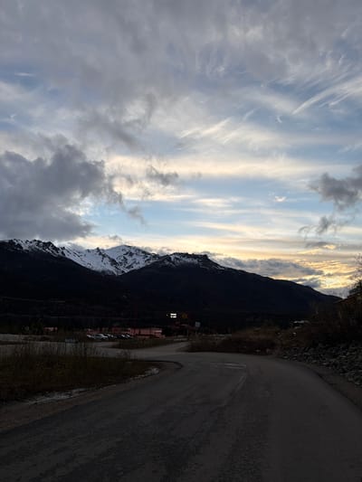 The image shows a landscape in Denali National Park and Preserve, United States. The scene includes a road to the right, a dark mountain range, partially covered in snow. The sky exhibits a mix of clouds, some dark and heavy on the left, with bands of light and a hint of sunset or sunrise towards the center. Buildings and parked vehicles are visible at the base of the mountain. The setting appears to be a mountainous area near a town or village.