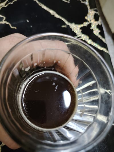 A close-up, overhead shot shows a glass of dark liquid, likely coffee or beer, sitting on a black, glossy surface with a swirling, marble-like pattern. The liquid has a thin layer of foam around the rim, with a few small bubbles scattered on the surface. The glass has a ribbed texture on its exterior. A person's hand, slightly out of focus, is visible on the left side, holding the glass. The lighting creates reflections on the glass and the surface, highlighting the glossy nature of the background. The scene is domestic and appears to be indoors, with no discernible features to place it in a specific city or geographical location.