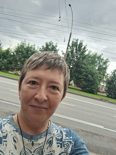 A medium-shot selfie shows a woman with short, graying hair and brown eyes looking directly at the camera. She is wearing a blue and white patterned shirt with a gold chain necklace. The background features a street with a lamppost and trees under a cloudy sky. The woman appears to be outdoors in Gorka-Pokrovskaya, Russia.