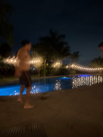 The image captures a nighttime scene around a swimming pool, characterized by significant motion blur, suggesting either fast movement or a long exposure. The setting is likely an outdoor area, possibly a resort or private residence, in Santa Ana Yancuitlalpan (Chilhuacán), Mexico.
The primary subject is a person, heavily blurred, appearing to be in mid-air or just entering the water. The person is wearing dark swimming trunks with a lighter stripe and is shirtless. Their body is stretched horizontally across the frame, with limbs extended. Another person's head or shoulder is partially visible at the top edge of the frame.
The scene features a blue-lit swimming pool, which takes up a significant portion of the right side of the image. The water shows wavy reflections of the lights. Along the edge of the pool and extending further back are decorative string lights, emitting warm yellow/white light, which are also blurred into streaks. These lights illuminate the surrounding area and create reflections on the water surface.
Behind the string lights and the pool, dark foliage, likely palm trees, is visible against a dark, nocturnal sky. The trees are also somewhat blurred but their general shape is discernible. To the right of the pool is a concrete or tiled surface. The overall impression is one of dynamic activity and playful movement in a festive, dimly lit environment. No visible text is present in the image.