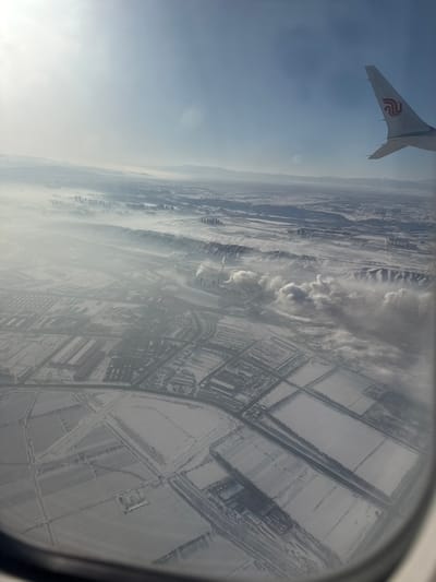 The image provides an aerial view of a snow-covered landscape observed from an aircraft window, with a section of a white airplane wing featuring a red phoenix logo visible in the upper right. The scene depicts the surroundings of Ürümqi, China, during winter.

In the foreground and lower midground, the terrain consists of extensive snow-covered fields, organized into rectangular plots by visible roadways or paths, interspersed with scattered low-rise industrial or warehouse structures. Towards the center of the midground, a large industrial complex is prominent, featuring multiple buildings, cooling towers, and several tall smokestacks actively emitting substantial white plumes of smoke or steam. These emissions create distinct, localized cloud formations and contribute to a widespread atmospheric haze that partially obscures distant features.

Behind the industrial complex, a distinctive, dark, elongated ridge or embankment, also lightly covered in snow, stretches horizontally across the scene. Further beyond this ridge, through the atmospheric haze, a dense urban area is discernible, characterized by numerous high-rise buildings. In the far background, snow-covered mountains define the horizon, partially obscured by the pervasive haze. The sky above is clear and light blue, with a bright sunlight glare present in the upper left quadrant. The environment is uniformly blanketed in snow, indicating a cold, winter season, with notable industrial activity impacting air visibility. No individuals are visible.