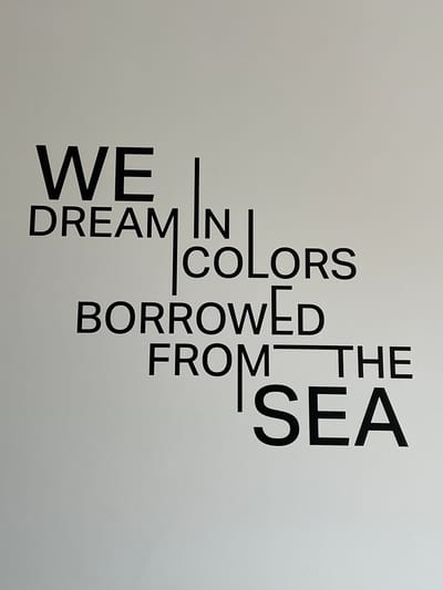 The image shows a stark white wall with black text arranged in a unique font. The text reads "WE DREAM IN COLORS BORROWED FROM THE SEA." The words are spaced out and some are stacked on top of each other, creating a visually interesting composition. The lighting is even, suggesting an indoor setting, possibly a gallery, cafe, or public space. The environment appears clean and minimalist. There are no people or specific landmarks visible in the image to indicate a location within Ras Al-Khaimah, United Arab Emirates.