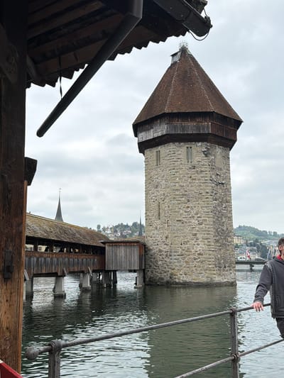 The image shows the Chapel Bridge and Water Tower in Lucerne, Switzerland, viewed from within the bridge structure. The stone water tower rises from the lake. The bridge's wooden roof and rafters frame the view, with a drainpipe prominently visible. Part of the bridge's metal railing and a person wearing a grey jacket are seen on the right. In the background, other buildings and the shoreline are visible. The sky is overcast.