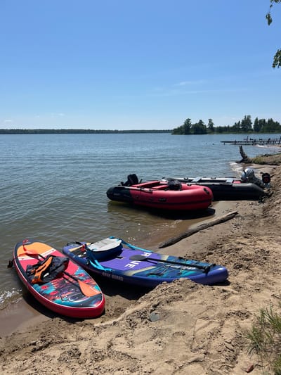 On a sandy shore of a lake, two inflatable paddleboards are resting. The paddleboard in the foreground is red with an orange paddle and a black and orange life jacket. The paddleboard behind it is purple with blue accents and appears to be labeled "RAVE BOARD." In the water behind the paddleboards are two inflatable boats. The boat in the foreground is red, and the boat behind it is dark green and has an outboard motor. In the distance, a tree line borders the lake, with a clear blue sky above. A small wooden dock is visible in the far right of the image.