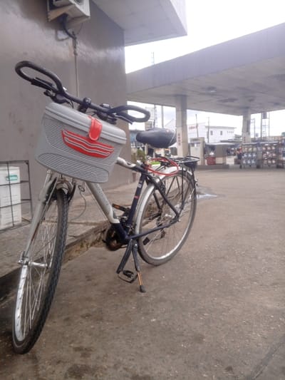 A light gray and dark blue bicycle is positioned upright on a gray concrete surface, supported by its kickstand, under the canopy of a petrol station. The bicycle features a large, light gray plastic front basket with red trim and wave-like decals, black handlebars, a black cushioned saddle, and a black rear rack. A red cable lock is secured around the saddle post. A white "CROSSWAVE" label is visible on the downtube. To the immediate left, an external electrical unit is mounted on a light gray wall. The background reveals additional elements of the petrol station, including fuel dispensing units, a visible red "UBA" sign on a support column, and a display rack holding multiple packaged items. Distant white buildings are visible beneath an overcast sky. The scene is located in Ajamimogha, Nigeria. No individuals are present in the image.
