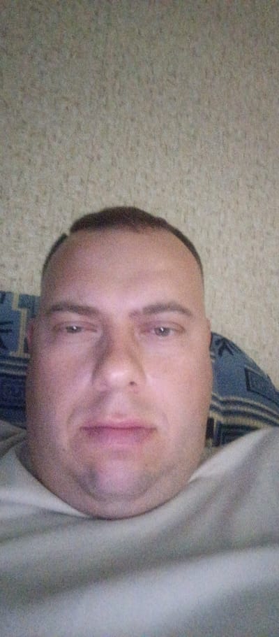 A close-up selfie of a man with short brown hair and a light complexion is captured against a textured, light-colored background. The man is looking towards the camera with his mouth slightly open. He is wearing a light-colored shirt. The lighting in the image appears to be artificial.