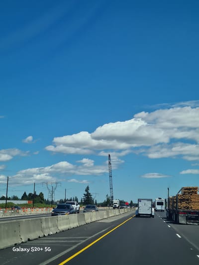 In Aurora, United States, on a bright, sunny day, a highway scene unfolds under a vast, clear blue sky dotted with fluffy white clouds. The perspective is from a vehicle traveling on the right lane of a multi-lane highway. Concrete barriers separate the lanes, and construction appears to be in progress, indicated by orange construction cones and more concrete barriers lining the side of the road. Power lines and transmission towers are visible in the distance, along with a line of trees and greenery. Several vehicles are on the road: a dark-colored sedan in the left lane, followed by a white pickup truck. Further ahead, a large truck is transporting lumber, and behind it, a recreational vehicle and a white trailer are also traveling. A tall construction crane stands prominently on the left side of the highway, near some trees. The road has yellow and white lane markings. In the lower left corner of the image, the text "Galaxy S20+ 5G" is watermarked.