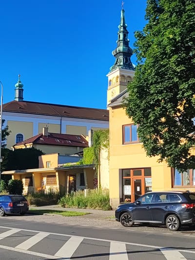 The image depicts a street scene in Nový Jičín, Czechia, on a sunny day with a clear blue sky. The foreground shows a crosswalk on a paved road, with two cars parked on the side. The car on the left is a dark blue station wagon, and the car on the right is a dark blue SUV.
Behind the cars, there is a yellow building with several windows. A large tree with green leaves partially obscures the view of the building and a church tower in the background. The church tower has a green spire and a clock face. The main body of the church appears to be light yellow with a brown roof. Another smaller building with a red tiled roof is situated in front of the church. The environment appears urban, with signs of vegetation growing along the side of the road and on some of the buildings.