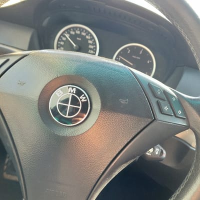 The image displays the interior of a vehicle, primarily focusing on a black, textured steering wheel and a portion of the instrument cluster. The steering wheel prominently features a circular, silver-rimmed BMW logo in its center, which exhibits a faint reflection of the surroundings. On the right spoke of the steering wheel, a set of four diamond-shaped buttons, arranged in a two-by-two grid, is visible. Below the right side of the steering wheel, a control stalk, possibly for cruise control or turn signals, is partially discernible. Behind the steering wheel, two circular analog gauges are visible. The left gauge, a speedometer, indicates a speed of approximately 50 km/h, with markings extending to 220 km/h. The right gauge, likely a tachometer, displays its needle at the zero position, with markings up to 4 (presumably x1000 RPM). No individuals are present in the frame, and no specific actions or interactions are depicted. The lighting suggests an ambient light source from outside the vehicle. The image provides no visual information to confirm the location as the city of Zlokuchene, Bulgaria.