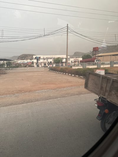 The image shows a view from inside a vehicle, looking towards the roadside in Iddah, Nigeria. A portion of the vehicle's interior is visible in the upper right corner, obstructing the view. A motorcycle with a carrier attached is partially visible in the upper right. The road is paved and gray. To the left of the road is a dirt verge, followed by buildings with the name "DEEPSTREAM OIL LTD" clearly visible on the facade. A mountain is visible in the background. Power lines are suspended above the scene. The sky is overcast and hazy.