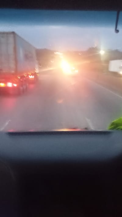 View from inside a vehicle, likely a truck, at dusk or dawn. A large semi-trailer is visible on the left, with its tail lights illuminated. Other vehicles are ahead, their headlights creating intense glare. The road appears wet. The dashboard is prominent in the foreground, with the sky just visible above the windshield. The overall scene is blurry and washed out due to the light and potentially a dirty windshield.