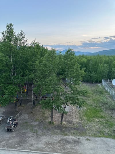 A wide shot captures a rural landscape at dusk in Sinegorye, Russia. The foreground features a dirt road with an all-terrain vehicle (ATV) parked on it. Behind the ATV, a cluster of tall birch trees with lush green foliage dominates the scene, partially obscuring a playground structure. Beyond the trees, a dense forest stretches into the distance, meeting a range of hazy blue mountains under a pale, cloudy sky. To the right, a white metal fence encloses a small area with a satellite dish. The ground is a mix of dirt, grass, and fallen leaves.