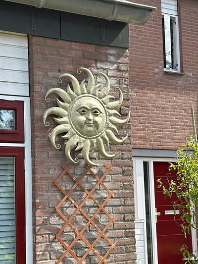 The image depicts an exterior wall of a brick building in Dronten, Netherlands. A decorative metal sun with a face is mounted on the wall. To the right of the sun is a wooden trellis. Above the brick wall are windows with white frames. Below the brick wall is a window with red frames. The scene appears to be outdoors during daylight. The environment consists of a residential building exterior in a Dutch city.