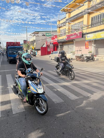 This is a photograph of a street scene in Dagupan, Philippines, likely captured during daylight given the strong shadows. In the foreground, a woman wearing a teal helmet and a black t-shirt with "NIKE" printed on it is riding a blue and black motorcycle. She is wearing ripped blue jeans and pink sandals. Her motorcycle is positioned on a pedestrian crossing. Behind her, another motorcycle is visible, ridden by a person wearing a black helmet and a black t-shirt. This motorcycle has a black storage box attached to the rear.
Further in the background, a blue truck is visible, loaded with red Coca-Cola crates. The street is lined with buildings, including a prominent yellow building with multiple windows and awnings, and other businesses with various signs, one of which reads "MOTOPOSH". White decorative flags or pennants are strung across the street and between buildings. The sky is blue with scattered clouds. The overall impression is a busy urban street with active traffic.
