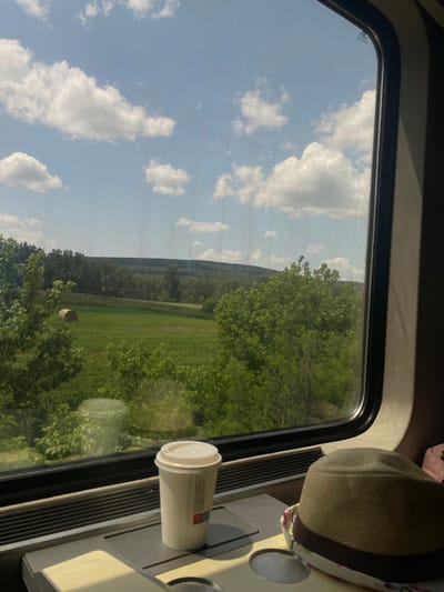 The image shows a view from inside a train, looking out the window at a green field with a hay bale in the distance. The window is in the upper right corner of the image. The train is traveling through the countryside, with a clear view of the sky and clouds. There is a cup of coffee and a hat on the table in the lower right corner of the image. The coordinates 47.566, 18.416 place the location of the scene in the city of Tatabánya, Hungary.