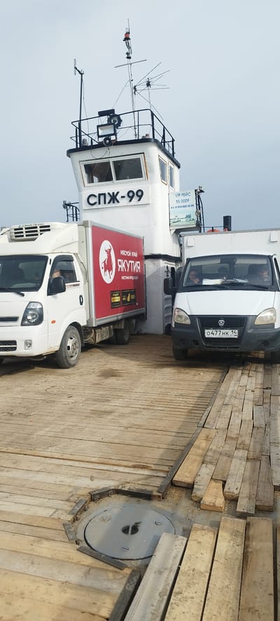The image shows two white delivery trucks parked on a wooden platform. Behind the trucks is a ferry with the designation "СПЖ-99" painted on its white bridge. The truck on the left has a red sign with a bull and the inscription "ЯКУТИЯ - МЯСНОЙ КРАЙ МЕСТНАА ПРОДУКЦИЯ" written in Russian. The truck on the right has a license plate reading "O477 HK 14". The platform appears to be made of rough wooden planks and features a circular metal hatch. The overall environment suggests a transport or loading area. Based on the text on the truck on the left, the location of the photo is likely Bestyakh, Russia.