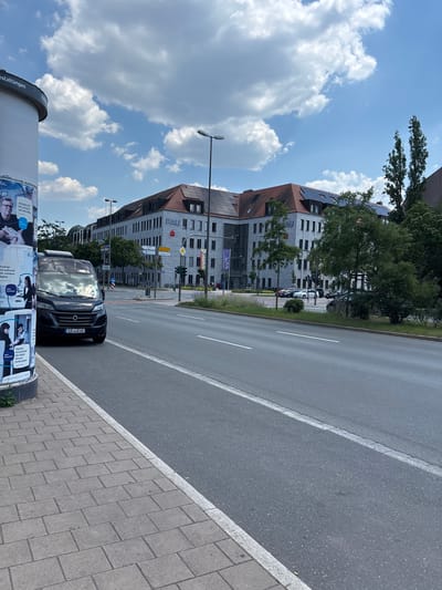 This is a vertical, medium-shot photograph taken outdoors in Erlangen, Germany. The scene depicts a street with a large building on the left and a road on the right. The sky is bright blue with scattered white clouds, suggesting it is daytime, likely late morning or early afternoon. The weather appears sunny.
On the left side of the image, a modern building with a reddish-brown tiled roof and a grey facade is visible. The building has many windows and solar panels on the roof. Text on the building reads "FAU," which likely refers to Friedrich-Alexander-Universität Erlangen-Nürnberg. In front of the building, trees and some parked cars can be seen. A light pole with two lamps stands in front of the building. At the very bottom of the left side, a large cylindrical structure, possibly a pillar for a billboard, is present with posters attached to it. Some of the text on the posters is legible, including "staltüngen," "Alltagstrot," "Wir arbeiten mit Menschen," "Analoge Verwaltung? Wir arbeiten digital," and "Digitale Verwaltung? Wir lieben Herausforderungen."
The right side of the image shows a paved road with a white line marking the lane. A dark-colored van or minivan is parked on the shoulder of the road, with its license plate visible as "ER-LD 16." The sidewalk next to the road is paved with grey rectangular bricks. The overall impression is a snapshot of an urban street scene on a clear day. There are no people prominently featured, but the posters suggest a public space with advertisements or information being displayed. The presence of the university building and the German license plate confirm the location context.