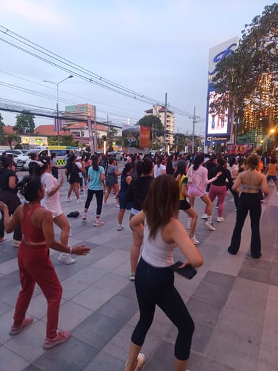 An outdoor aerobics class is taking place in Hua Hin City Municipality, Thailand, on a plaza paved with square tiles. The participants, mostly women, are dressed in sportswear and sneakers, and are engaged in synchronized movements. They are facing away from the viewer towards a brightly lit commercial area. A large digital billboard displays images of people. Behind the participants are cars, a Thai flag, and buildings, suggesting an urban setting. Overhead, there are electrical wires. The sky is a pale dusk color.
