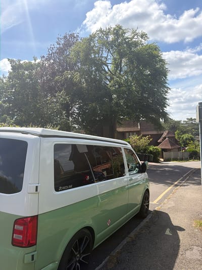 The image captures a street scene in Guildford, United Kingdom, featuring a two-tone light green and white Volkswagen Transporter parked alongside a road. The road is marked with a yellow line along the curb. The vehicle's side is prominently displayed, including details like the "REDLINE" logo on a window. A large tree dominates the left side of the frame, partially obscuring a house in the background. The sky is visible above the tree, featuring scattered clouds and bright sunshine. A shadow is cast on the road near the vehicle, and portions of a building can be observed behind the tree and the houses.