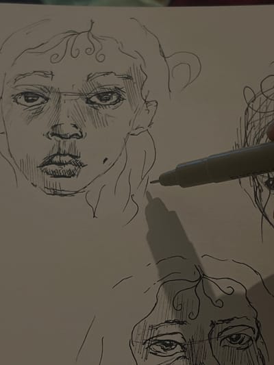 An overhead shot captures a sheet of plain white paper displaying multiple ink drawings of human faces. The central and most complete drawing depicts a stylized face with wide, darkly defined eyes, a pronounced nose, and full lips detailed with vertical lines. The forehead features distinct wavy, scroll-like patterns, and hatching provides contour on the cheeks and nose. A small circular mark is present on the right cheek. Partially visible in the bottom-right corner is another drawing of a similar face, showing the eyes and forehead with comparable swirling patterns and hatching. A gray fine-tipped pen is held by human fingers (thumb and index finger visible) over the paper in the mid-right section, with its tip poised near the lower drawing. A prominent shadow of the pen and hand is cast onto the paper, indicating an external light source. The background surrounding the paper is dark and indistinct. This scene is located in the city of Nor Kharberd, Armenia.