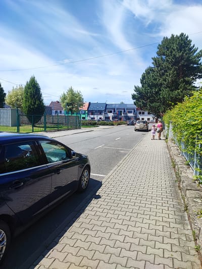 The image depicts a suburban street scene in Nový Jičín, Czechia, on a bright, sunny day with scattered clouds in a blue sky. In the foreground, a dark blue sedan is parked on the left side of the road, casting a shadow on the grey, interlocking paver sidewalk. The sidewalk runs along the right side of the street, bordered by a low stone wall and a vibrant green hedge.
Further down the sidewalk, two people, a woman and a young girl, are walking away from the camera. The woman is wearing a pink tank top and shorts, while the girl has on a purple dress and hat. They are walking on the paver sidewalk towards a row of modern houses with colorful facades and dark tiled roofs, which line the street in the background.
Across the street, another car, a beige SUV, is parked. The street itself is asphalt with visible white lane markings. A green metal fence separates the sidewalk area from a grassy area with trees behind it. A tall evergreen tree stands prominently on the right side of the image, behind the hedge and near the houses. The overall impression is one of a peaceful, residential neighborhood during daytime. There is no visible text in the image.