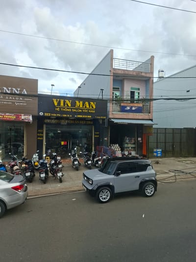 The image shows a street scene in B'Lao, Vietnam. A light gray SUV is parked on the right side of the street, facing left. In front of it, parked on the sidewalk, are several motorcycles. Behind the motorcycles are storefronts. The central storefront has a large black sign with yellow lettering that reads "VIN MAN" and "HỆ THỐNG SALON TÓC NAM". Below this, it says "323 Trần Phú, Lộc Sơn" and a phone number, "0966.919.856". The entrance to the salon is visible, showing a well-lit interior. To the left of the "VIN MAN" salon is another storefront with a brown sign that says "ANNA R SALON". To the right of the "VIN MAN" salon, a taller building is visible with a pink and light blue facade. Electrical wires are strung across the top of the image, and a water tank is on the roof of one of the buildings. The sky is overcast.