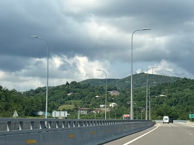 The image depicts a highway overpass in the region of Ajara, Georgia, under a cloudy sky. In the foreground, a concrete barrier with yellow markings runs along the edge of the road. Several modern streetlights with curved arms are visible, lining the highway. Beyond the barrier, a lush, green, tree-covered hillside is dotted with scattered houses and buildings. The road curves gently to the right, with a white van and a black car visible in the distance, driving away from the camera. A green road sign with white lettering is also present on the right side of the road. The overall scene suggests a natural, somewhat rural landscape with infrastructure for transportation.