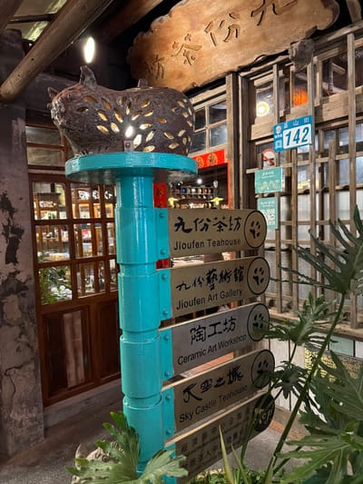 The image depicts a street scene in Jiufen, Ruifang District, New Taipei City, Taiwan, at night. A teal-painted, multi-segment pole stands prominently in the foreground. Atop the pole is a rustic, illuminated metal lantern shaped like an animal, featuring numerous leaf-shaped perforations through which light emanates. Vertically mounted on the pole are four rectangular signs, each displaying an establishment name in both English and Chinese (Traditional), accompanied by a paw print icon: "Jiufen Teahouse" (九份茶坊), "Jiufen Art Gallery" (九份藝術館), "Ceramic Art Workshop" (陶工坊), and "Sky Castle Teahouse" (天空之城). Partially obscured smaller signs are visible beneath these.
To the left of the pole, a building entrance with dark wooden-framed glass doors and windows reveals a warmly lit interior featuring shelves and various items. To the right, a traditional wooden facade with multiple window panes displays a blue and white street sign reading "基山街 142" (Jishan Street 142) and a green sign indicating "Business Hours 11:00-20:00". A large, carved wooden sign with Chinese characters is affixed above this facade. Large, green, broad-leaf plants are visible in the lower left and right portions of the frame. The scene features no visible human subjects, presenting a commercial or cultural area with traditional Taiwanese architectural elements.