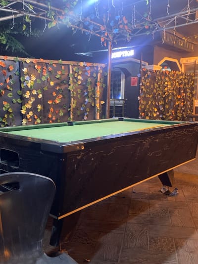The image displays an unoccupied outdoor or semi-covered entertainment area at night in Igbessa, Nigeria. A black pool table with a green felt playing surface is centrally positioned, with a single cue stick resting on its far edge. The table's dark exterior shows visible scuffs and wear. A portion of a dark grey plastic chair is visible in the lower-left foreground. The ground is paved with brown, textured tiles, with small pieces of scattered debris visible beneath the pool table. Behind the table, a dark wall or fence structure is adorned with artificial foliage, featuring predominantly autumn-toned leaves in shades of orange, green, and light blue. Above the scene, a metal trellis supports artificial vines and illuminated string lights. To the right, a dark building entrance is visible, featuring a partially illuminated sign displaying "SCRIPTION," indicating a "RECEPTION" area. No individuals are present.