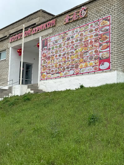 The image shows the exterior of a Chinese restaurant named "ЗОЛОТОЙ ДРАКОН" (Golden Dragon) in Chuguyevka, Russia. The building is made of brick. A large banner displaying various Chinese dishes is mounted on the exterior wall. There is a glass door entrance with a set of stairs leading up to it. Red lanterns are hanging near the entrance. The restaurant is situated on a grassy slope.