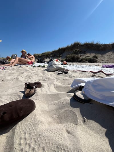 The image depicts a sunlit beach scene on a clear day in Skagen, Denmark, characterized by light-colored sand and a backdrop of natural dunes. In the immediate foreground, textured sand displays a distinct hexagonal tire tread pattern on the lower left. Scattered personal effects include a dark brown suede clog and a brown two-strapped sandal on the left, and a white garment draped over a dark-soled flip-flop on the right.

Further back, two individuals are reclined on beach blankets. The person on the left wears a straw hat and sunglasses, with legs extended. The individual on the right is partially visible, wearing a yellow bikini top. Both appear to be relaxing on the sand. Adjacent to them are various items: a light-colored tote bag with the visible text "ABB National Electr", a small orange and white patterned ball, a pair of dark-framed glasses, and dark flip-flops. Red packaging is also present on the sand near the individuals. The background features a line of natural sand dunes topped with sparse, dry grasses, extending under a clear blue sky. Shadows indicate strong overhead sunlight.