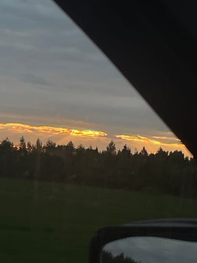 The image depicts a cloudy sunset over a tree line in Povarenki, Russia. The sky is a mix of gray clouds with streaks of orange and yellow light breaking through, suggesting the sun is low on the horizon. A dark silhouette of a forest borders the horizon, with the tops of the trees visible against the sky. In the foreground, a dark, curved object, likely the frame of a vehicle or building, partially obstructs the view. The lower part of the image shows a dark green grassy area. No people or specific activities are visible.