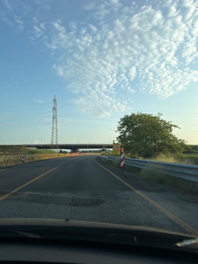 The image depicts a rural road in Novara, Italy, with a clear sky and scattered clouds. The road is asphalt with yellow lane markings. On the left side of the road, there are warning cones and bollards, and a tall electricity pylon. In the background, an elevated highway overpass spans the scene. On the right side of the road, a guardrail separates the road from overgrown vegetation and a large, leafy tree. A speed limit sign indicating 30 km/h is visible on the right. The view is from the perspective of someone inside a vehicle, looking forward through the windshield.