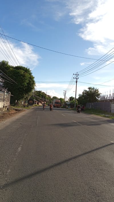 The image depicts a street scene in Cotbadatunong, Indonesia, under a clear blue sky with scattered white clouds. The perspective is from ground level, looking down a paved road. On the left side of the road, a large, leafy green tree stands beside a fence. Further down the road, there are several buildings and a large circular sign with the word "AREA" in it. A few motorcycles are visible on the road, with riders. In the distance, a large orange truck is driving on the road, and on the right side, there are more buildings, including some under construction with scaffolding. Electrical poles and wires are strung across the sky, a common sight in urban and semi-urban environments in Indonesia. The overall impression is of a typical day in a bustling Indonesian town.