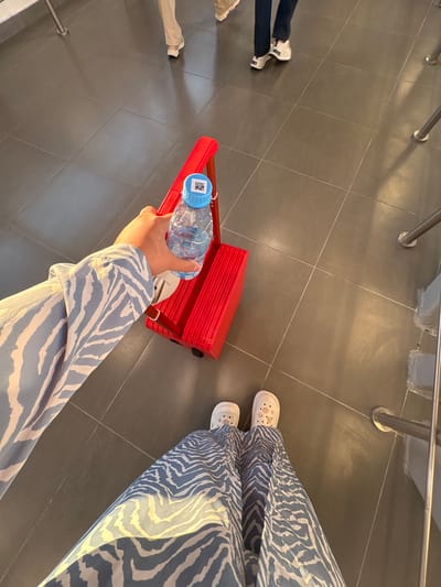 In a high-angle, close-up shot of feet and a hand on a tiled floor, a person wearing a blue and white zebra-print maxi dress and white clogs decorated with shell-like patterns stands on dark gray square tiles. The person's left hand, adorned with a blue and white zebra-print long-sleeved shirt, holds a clear plastic water bottle with a blue cap. The bottle features a QR code on its label. The bottle is positioned above a small, bright red rolling suitcase with a silver handle. The background shows more of the dark gray tiled floor and appears to be an indoor public space, possibly a waiting area or hallway. In the mid-ground, two other individuals are visible from the knees down, one wearing beige pants and white sneakers, and the other wearing dark blue pants and white sneakers. Metal railings and supports are visible on the right side of the frame. The location is identified as being in Matveevka, Russia.