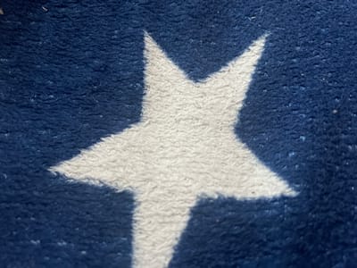 A close-up image of a textured fabric with a repeating pattern of white stars on a dark blue background. The fabric appears to be soft, possibly a fleece or similar material. The lighting is even, highlighting the texture of the fabric. There are no people, actions, or interactions visible. The location cannot be determined from the image.