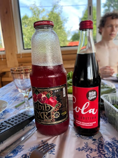 The image shows a table set outdoors on a sunny day, with a person in the background. On the table, there are two bottles of drinks, a glass, a remote control, and cutlery. One bottle is filled with a red liquid, identified as "Habib NAR Pomegranate juice drink" with the text "Azeri-Grand" on the cap. The other bottle contains a dark brown liquid, labeled "Cola LIMITED EDITION" with a logo featuring a crest and the text "Бавария" in Russian. The background suggests a rural or suburban setting with trees and a blue sky visible through a window or open structure. The person in the background appears to be a young male, shirtless, with curly hair. The table is covered with a patterned tablecloth. The overall scene depicts a casual outdoor gathering.