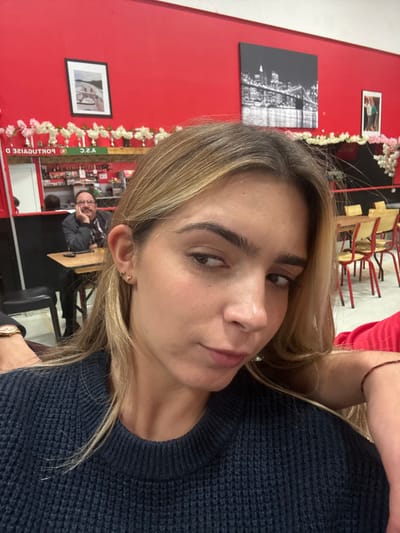 The image displays a young woman in the foreground, taking a selfie inside a casual restaurant or bar. She has light brown hair, is wearing a dark blue ribbed sweater, and exhibits a subtle expression looking towards her right (viewer's left). Two small stud earrings are visible in her right ear. A portion of a left arm wearing a watch is visible on the far left, and a right arm with a thin dark bracelet is partially visible on the far right.
In the mid-ground, an adult male with glasses and a mustache is seated at a wooden table, resting his chin on his left hand. He is wearing a plaid shirt under a dark outer layer.
The background features vibrant red walls. A green sign, reflected in a mirror, reads "PORTUGAISE D. A.S.C.", indicating a Portuguese cultural or sports association, which places the establishment in Décines-Charpieu, France. Above this, a string of white artificial flowers is draped along a ledge. Wall decor includes a framed landscape photograph with a black border, a large black and white photograph of the Brooklyn Bridge, and a smaller framed image with two figures. Multiple wooden tables with red metal-framed chairs are arranged in the background on the right. A bar counter with various bottles is visible in the reflection on the left side of the background.