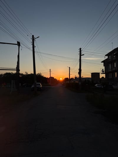 The image depicts a street scene in Petkovo, Bulgaria during sunset. The sun is low on the horizon, casting a warm glow across the sky and illuminating the clouds. The street is paved and appears to be cracked in places. There are power lines crisscrossing the sky, supported by utility poles. On the right side of the street, there is a multi-story building with balconies and satellite dishes. Several cars are parked along the street and in what appears to be a parking area. Trees and foliage line the sides of the road. In the foreground, on the left, a wire fence and soccer goalposts are visible. The overall impression is one of a quiet, residential street at the end of the day.