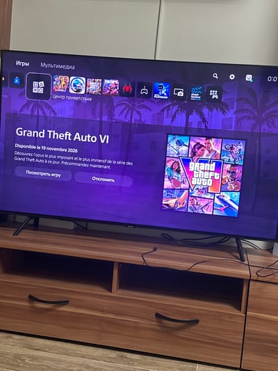 The image shows a television screen displaying the Grand Theft Auto VI game interface, with the title and release date prominently featured. The release date is indicated as November 19, 2026. Below the title, there is a description of the game in French, mentioning it as the most ambitious and immersive opus in the Grand Theft Auto series to date and encouraging pre-orders. Two buttons are visible: "Посмотреть игру" (View game) and "Отклонить" (Decline). The screen also shows a grid of images related to the game. The television is placed on a wooden media console. The background appears to be an indoor setting, possibly a living room, with a wall that is light-colored and appears to be a cabinet or closet. The overall scene is well-lit. There is no indication of the specific location within Villeurbanne, France.