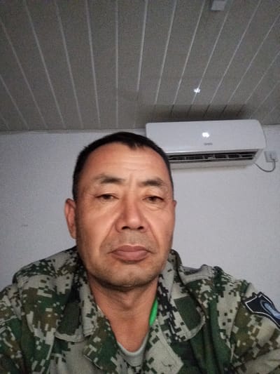 A medium shot portrait of a middle-aged East Asian man wearing a camouflage jacket is captured from a slightly low angle. The man's head is turned slightly to his right, and his gaze is directed towards the camera. He has short, dark hair, a prominent brow, and visible wrinkles around his eyes and forehead. His mouth is closed, and his expression is neutral. The man is wearing a green t-shirt underneath the camouflage jacket, which has a pixelated digital camouflage pattern in shades of green, gray, and black.
Behind the man, a white air conditioning unit is mounted on a light gray wall. The air conditioner has the brand name "DIVO" visible on its front panel, along with a digital display showing "05". The ceiling above the man is paneled with long, narrow strips, creating a ribbed texture. The overall environment appears to be indoors, possibly an office or residential space, given the presence of the air conditioner and the paneled ceiling. The lighting in the scene is somewhat dim, casting subtle shadows on the man's face. The image has a grainy texture, suggesting it was taken with a low-light capable camera or under low-light conditions. The provided location information suggests the photo was taken in the vicinity of the Түндүк айланып өтүүчү жол 84 in Kyrgyzstan, although the image itself does not contain any explicit geographical markers.