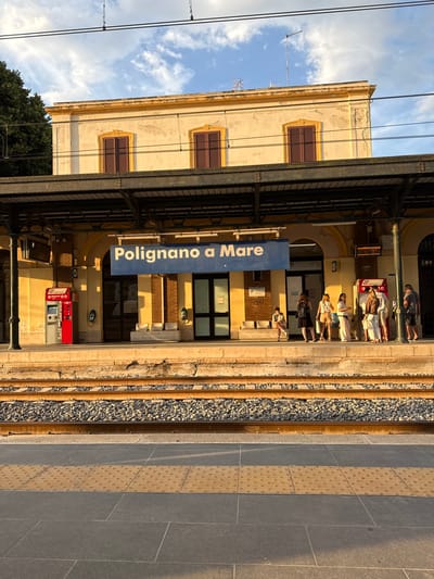 The image depicts the train station platform at Polignano a Mare, Italy, under clear, late afternoon lighting conditions. The primary subject is a two-story, pale yellow station building with a weathered facade and minimal architectural detail, featuring three upper-level windows with closed brown louvered shutters. Above the building, a blue sky with scattered white clouds is visible, along with overhead power lines for trains and two antenna structures on the roof.
Extending from the station building is a dark metal canopy that shelters the platform. Underneath this canopy, a prominent blue sign with white lettering displays "Polignano a Mare." The ground floor of the station building features arched openings, some appearing as entrances.
On the concrete platform, two "Trenitalia Biglietti Self Service" ticket machines are visible; one red and grey machine is on the left side, and another similar machine is on the right. A group of approximately seven adults is gathered near the right-hand ticket machine, standing and appearing to wait or interact. One individual is seated on a white bench to the left of this group, engaged with a mobile phone. Another empty white bench is positioned further left. The people are casually dressed in warm-weather attire.
In the immediate foreground, two parallel train tracks are laid on a bed of grey ballast. The tracks and ballast are illuminated by warm, low-angle sunlight, casting distinct shadows. The platform surface includes a section of yellow tactile paving with raised dots, indicating a warning strip. A dense green tree is partially visible on the far left, behind the station building.