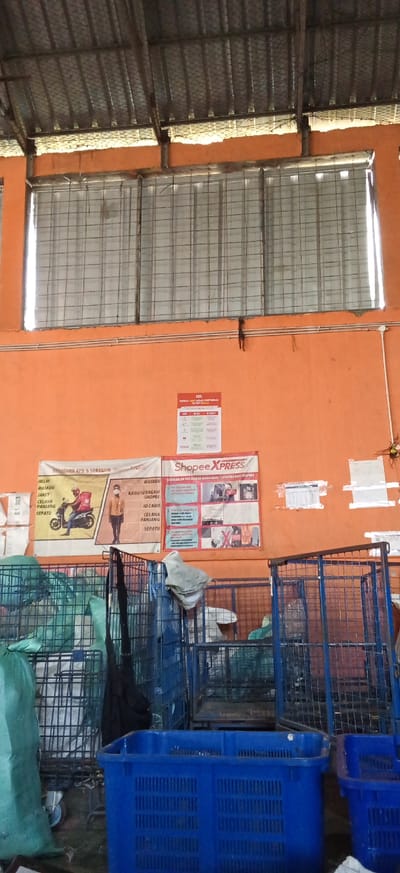 This image captures a cluttered interior space, likely a warehouse or storage area, with an orange-painted wall as the backdrop. In the upper section of the wall, a poster with red and white text is visible, along with a white poster on the left. Below these, two larger posters are displayed. The left poster features an illustration of a person on a motorcycle with a delivery bag and a list of items including "HELM", "MASKER", "JAKET", "CELANA PANJANG", and "SEPATU". The right poster is branded with "Shopee XPRESS" and contains text and images related to package handling and delivery, including depictions of goods and individuals.

The lower portion of the image is dominated by several industrial blue metal cages, some of which contain bags and bundles. In the foreground, a large blue plastic crate is prominent, with another smaller blue crate visible to its right. The floor appears to be concrete and is covered with various items, including green bags and a black garment. The ceiling is a corrugated metal structure with visible supports. The overall impression is one of a busy operational area focused on logistics and delivery services. The context of Kiangroke, Indonesia, suggests this could be a distribution hub or a storage facility for a company like Shopee.