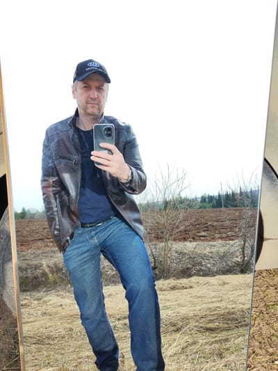 A middle-aged man is taking a selfie in a mirror, standing outdoors in a field. He wears a dark cap with the letters "ADA MOTORS" on it, a brown leather jacket over a navy blue shirt, blue jeans, and a belt. He is holding a smartphone with dual rear cameras. The background reflected in the mirror shows a rural landscape with brown fields, dry grass, bare trees, and a distant forest. The image was taken in Нечкино, Russia.
